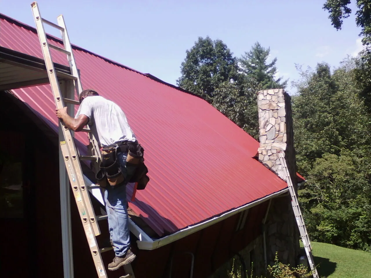 Roofer on a ladder beside a red metal roof and stone chimney — Soffit & Fascia Repair by Roofing Brothers in Arkport